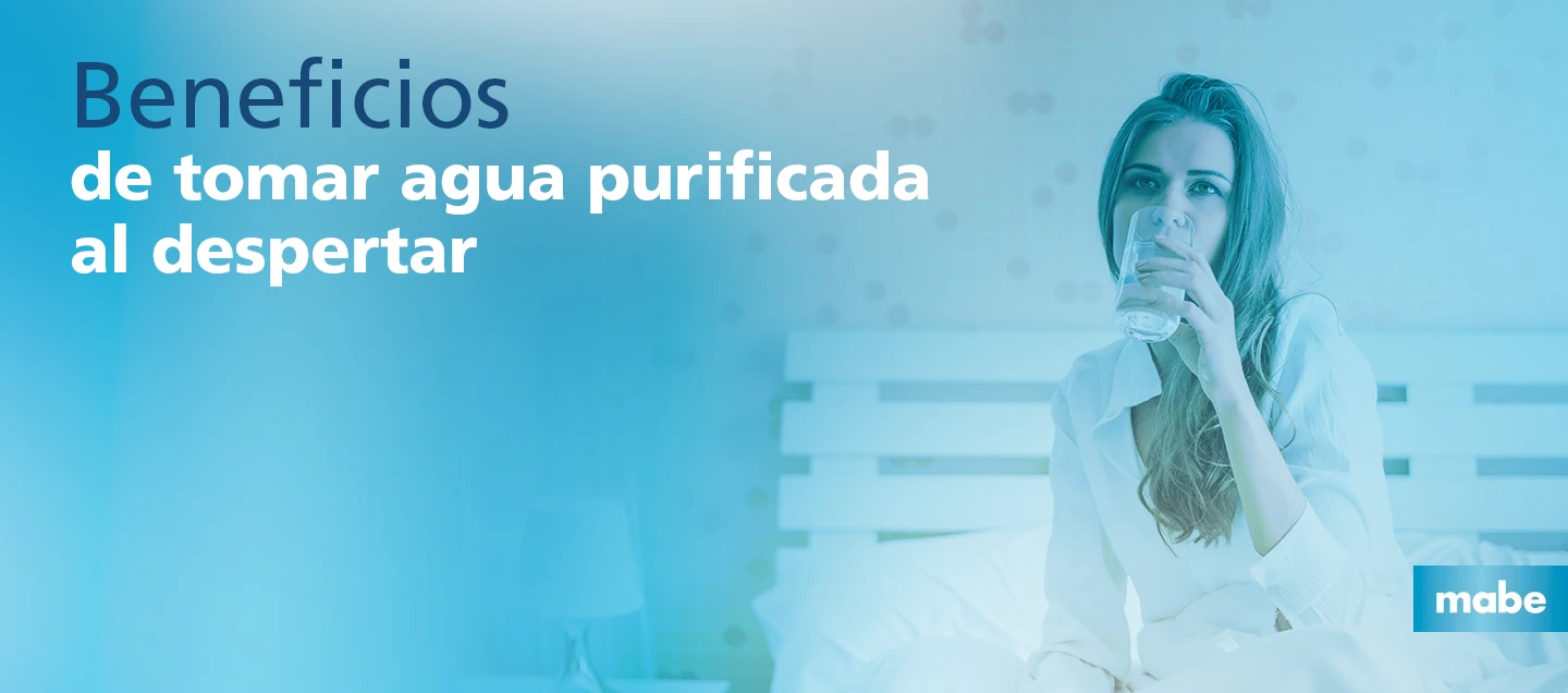 Mujer sentada en la cama al despertar, bebiendo un vaso de agua purificada, destacando los beneficios de comenzar el día hidratado.